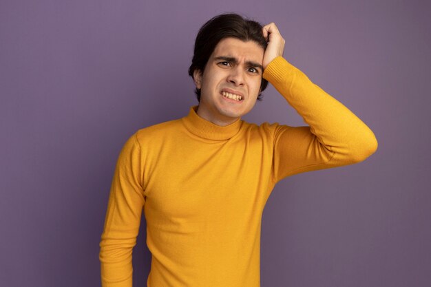 Preoccupato giovane bel ragazzo che indossa un maglione dolcevita giallo che mette la mano sul tempio isolato sulla parete viola