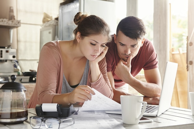 Preoccupato giovane coppia di sposi caucasica che legge una notifica importante dalla banca durante la gestione delle finanze nazionali e il calcolo delle spese al tavolo della cucina, utilizzando il computer portatile e la calcolatrice