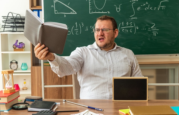 preoccupato giovane insegnante con gli occhiali seduto alla scrivania con materiale scolastico e mini lavagna su di esso in aula allungando il blocco note guardandolo