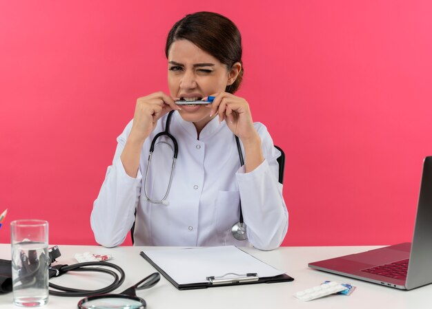 Preoccupato giovane medico femminile che indossa abito medico con lo stetoscopio seduto alla scrivania lavora sul computer con strumenti medici tenendo la penna in bocca sulla parete rosa isolata con spazio di copia