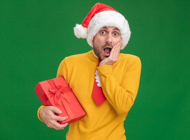 Preoccupato giovane uomo caucasico che indossa cappello di Natale e cravatta che tiene confezione regalo tenendo la mano sul viso isolato sulla parete verde con lo spazio della copia