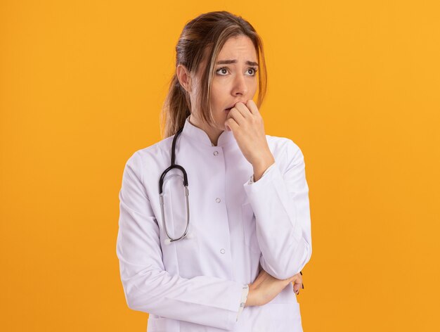 Preoccupato guardando il lato giovane medico femminile che indossa una veste medica con lo stetoscopio che mette la mano sulla bocca isolata sulla parete gialla