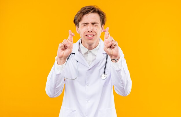 Preoccupato per gli occhi chiusi, giovane medico maschio che indossa un abito medico con lo stetoscopio che incrocia le dita isolate sulla parete arancione