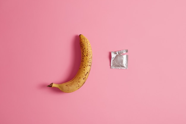 Preservativo disimballato e una banana intera su sfondo rosa studio.