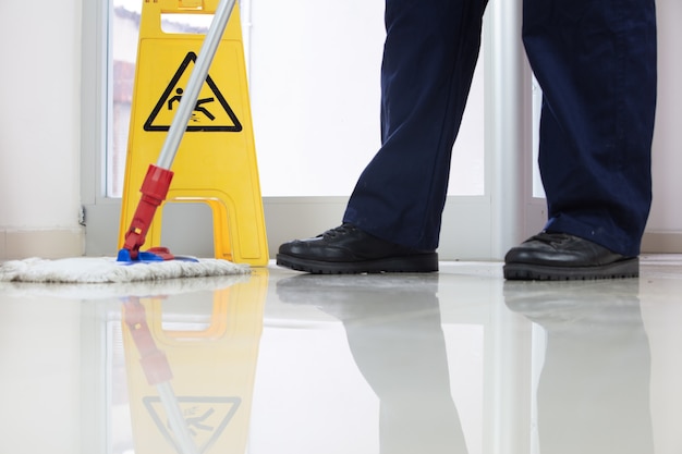 Primo piano ad angolo basso di una persona che pulisce il pavimento con una scopa vicino a un cartello giallo di attenzione sul pavimento bagnato wet