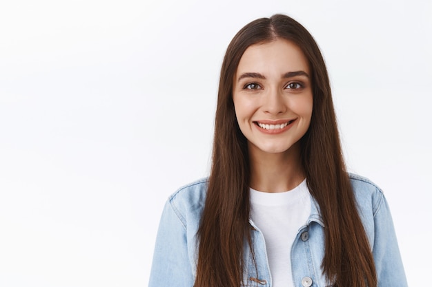 Primo piano attraente giovane donna tenera e femminile con acconciatura lunga, sorridente sciocco e carino con espressione casual rilassata, in piedi su sfondo bianco in giacca di jeans, mostra emozione positiva