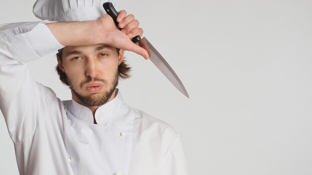 Primo piano chef maschio in uniforme che tiene la mano sulla testa che sembra stanco dopo una dura giornata di lavoro su sfondo bianco