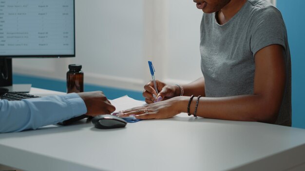 Primo piano dei documenti di controllo per la firma del paziente per l'esame in gabinetto. Giovane donna alla consultazione sanitaria che firma i documenti medici mentre parla con il medico del trattamento