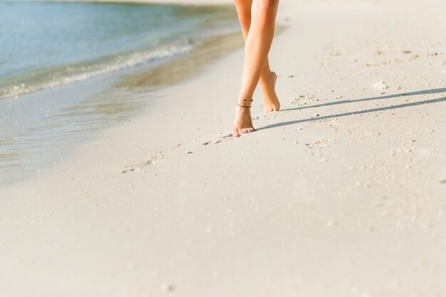 Primo piano dei piedi della ragazza sottile abbronzata nella sabbia. Cammina vicino all'acqua. La sabbia è oro