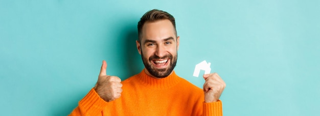 Primo piano del bene immobile dell'uomo soddisfatto che mostra pollice in su e piccola casa di carta maket che sorride sta compiaciuta