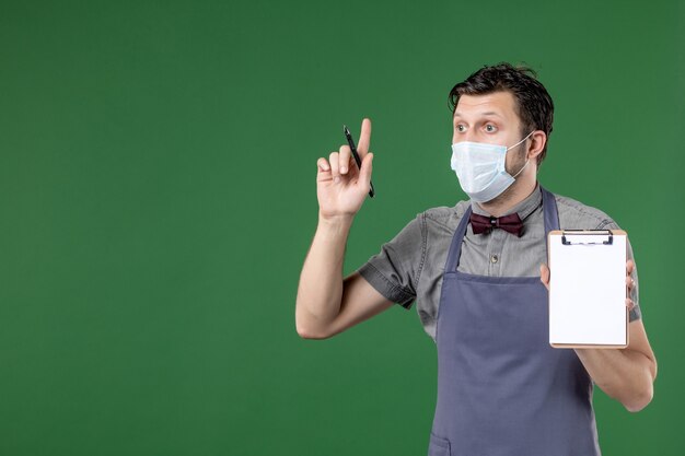 Primo piano del cameriere maschio concentrato in uniforme con maschera medica e tenendo la penna del libro degli ordini rivolta verso l'alto su sfondo verde