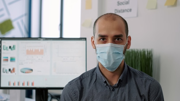 Primo piano del dipendente che indossa una maschera medica di protezione che guarda nella telecamera mentre si trova su chiar nel nuovo normale ufficio aziendale. Lavoratore nel rispetto del distanziamento sociale per evitare il contagio da covid19