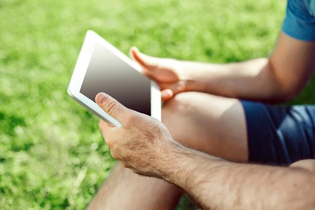 Primo piano del giovane vestito casual utilizzando la tavoletta digitale moderna mentre era seduto al parco, uomo hipster che collega il social networking all'esterno, luce del sole