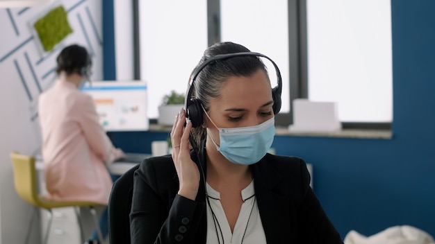 Primo piano del manager esecutivo con maschera facciale e auricolare che lavora su un computer portatile nell'ufficio dell'azienda durante la pandemia globale di coronavirus, . I colleghi mantengono il distanziamento sociale per prevenire la malattia da virus