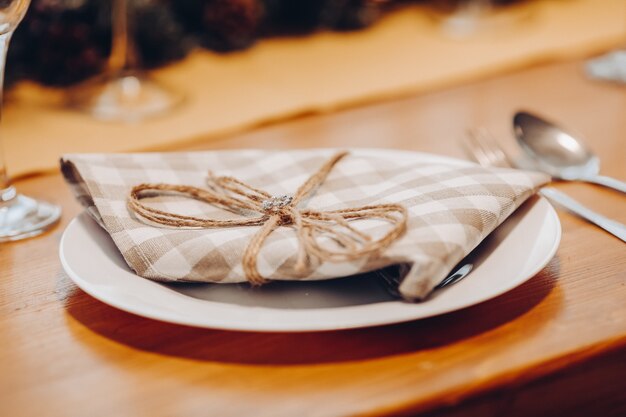 Primo piano del piatto in ceramica con tovagliolo controllato marrone e bianco con nastro sul tavolo da pranzo in legno con posate. Concetto di cena di Natale.