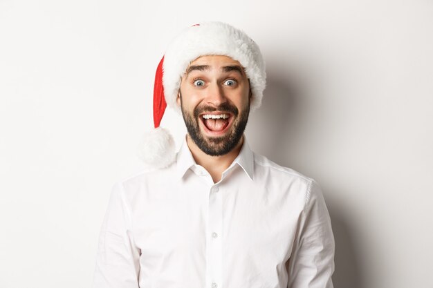 Primo piano del ragazzo barbuto felice in cappello della Santa, che sembra sorpreso, festeggia il Natale, in piedi