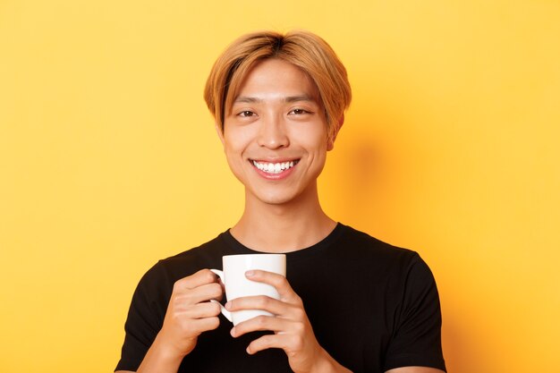 Primo piano del ragazzo sorridente asiatico soddisfatto felice, che tiene tazza con caffè, bevendo e guardando soddisfatto, in piedi sopra il muro giallo