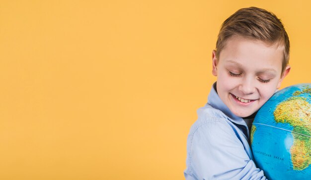 Primo piano del ragazzo sorridente che abbraccia globo contro fondo giallo