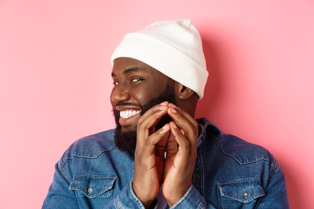 Primo piano del subdolo modello maschio afroamericano che ha un'idea, intriga qualcosa, dita del campanile e sorride sornione, in piedi su sfondo rosa.