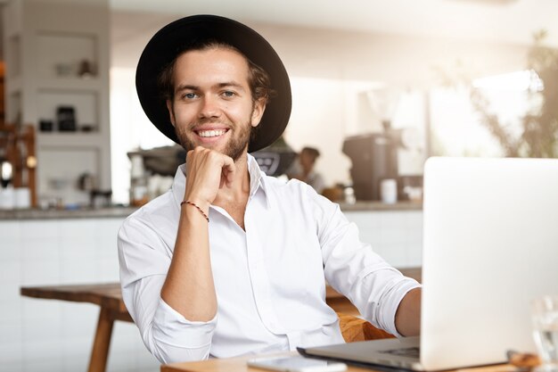 Primo piano del volto del giovane alla moda con il computer portatile, utilizzando la connessione internet ad alta velocità durante il pranzo nell'accogliente caffetteria interna.