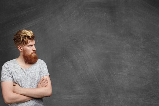 Primo piano del volto del giovane alla moda rossa con l'acconciatura alla moda e la barba folta vestito casualmente in piedi nell'angolo inferiore destro della lavagna vuota, tenendo le braccia incrociate e distogliendo lo sguardo