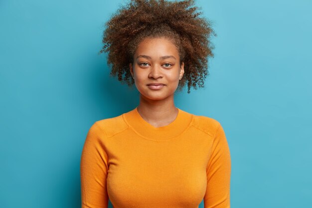Primo piano del volto della ragazza adolescente dai capelli ricci seria sembra soddisfatto vestito con un maglione arancione casual.