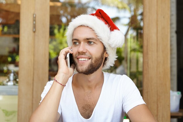 Primo piano del volto di giovane uomo con la barba lunga vestito con t-shirt bianca e cappello rosso di Babbo Natale che sembra felice mentre parla al cellulare con la sua ragazza, ascoltando le sue calorose congratulazioni per il Natale