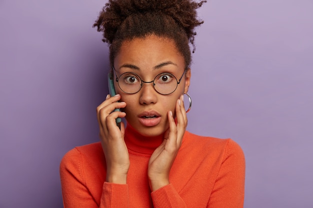 Primo piano del volto di una donna dalla pelle scura, emotiva, sbalordita, ansima dalla paura, sente notizie spiacevoli, conversa al telefono, indossa occhiali da vista e dolcevita, modelle su sfondo viola.