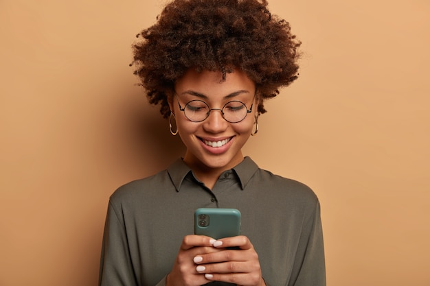 Primo piano del volto di una donna riccia che indossa occhiali trasparenti, camicia grigia, utilizza un'app online gratuita su smartphone, visualizza immagini, indossa occhiali rotondi e camicia