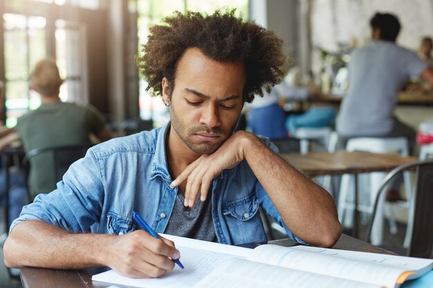 Primo piano del volto di uno studente laureato serio dalla pelle scura in camicia elegante blu che studia alla mensa o in uno spazio di coworking mentre si prepara per gli esami finali, prendendo appunti nel quaderno, avendo concentrato l'espressione