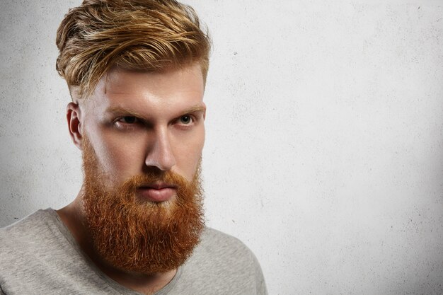 Primo piano del volto di uomo alla moda giovane hipster con barba folta e taglio di capelli alla moda che esamina la distanza con un'espressione seria e fiduciosa sul viso.