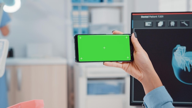 Primo piano dell'ortodontista che tiene lo schermo verde orizzontale sullo smartphone nell'ufficio per l'igiene orale. Dentista guardando il modello di mockup e lo sfondo isolato per la cura dei denti e l'odontoiatria.