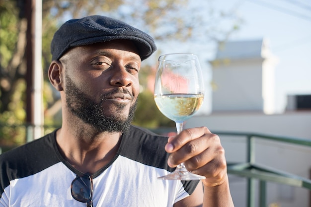 Primo piano dell'uomo africano felice che tiene un bicchiere di vino. Giovane bell'uomo in berretto nero che degusta vino bevendo guardando la fotocamera sullo sfondo della città e del cielo blu. Celebrazione, appuntamenti, concetto di momenti felici