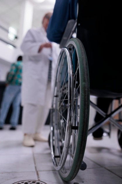 Primo piano dell'uomo anziano che vive con disabilità utilizzando la sedia a rotelle nella reception di una clinica privata che parla con il medico dell'appuntamento. Messa a fuoco selettiva sulla ruota della sedia a rotelle nella trafficata hall dell'ospedale.