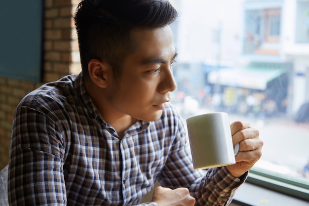 Primo piano dell'uomo asiatico che mangia il suo caffè di mattina alla finestra