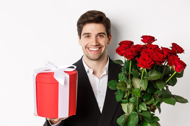 Primo piano dell'uomo barbuto bello in vestito, che tiene presente e mazzo di rose rosse, sorride alla macchina fotografica, in piedi su sfondo bianco