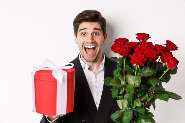 Primo piano dell'uomo barbuto bello in vestito, che tiene presente e mazzo di rose rosse, sorride alla macchina fotografica, in piedi su sfondo bianco