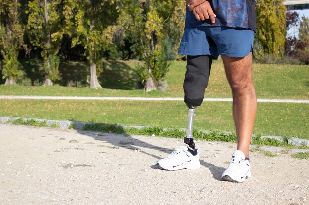 Primo piano dell'uomo caucasico con la gamba meccanica. Sportivo in pantaloncini blu e scarpe da ginnastica bianche fotografato nel parco. Sport, concetto di disabilità