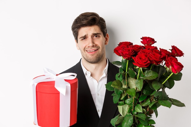 Primo piano dell'uomo scettico in tuta, con in mano un mazzo di rose rosse e un regalo, riluttante in piedi contro uno sfondo bianco.
