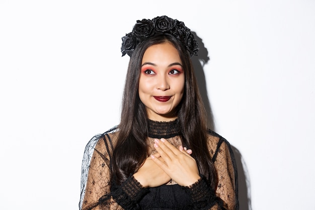 Primo piano della donna asiatica sorridente nostalgica che guarda sognante nell'angolo in alto a sinistra, tenendo le mani sul cuore, indossando abiti di pizzo gotico per la festa di Halloween