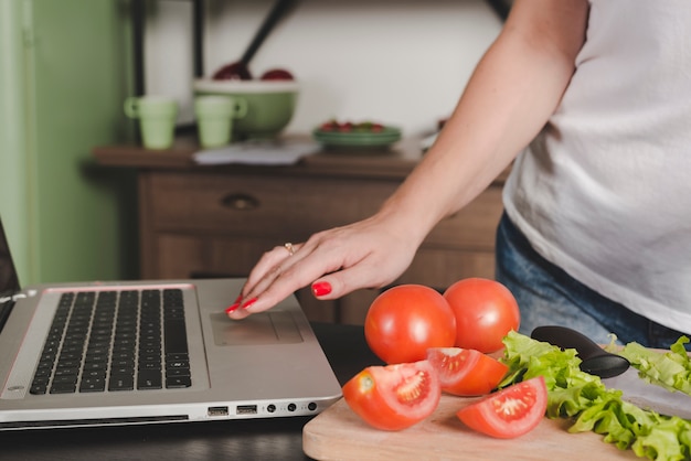 Primo piano della donna che utilizza computer portatile con i pomodori e la lattuga sul contatore di cucina