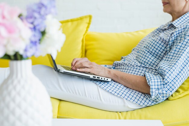 Primo piano della donna senior che si siede sul sofà che scrive sul computer portatile