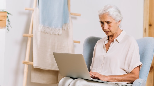 Primo piano della donna senior che si siede sulla sedia facendo uso del computer portatile