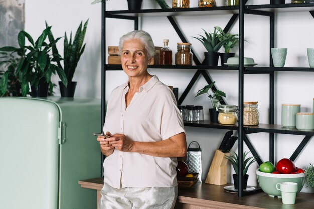 Primo piano della donna senior sorridente che utilizza condizione mobile nella cucina