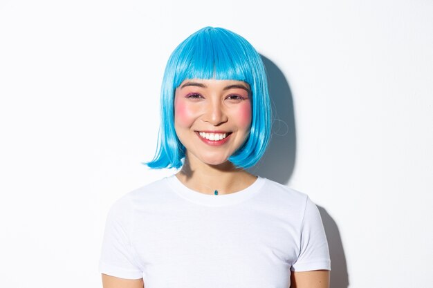 Primo piano della femmina asiatica sveglia che celebra Halloween in parrucca blu, sorridendo felice alla macchina fotografica, in piedi.