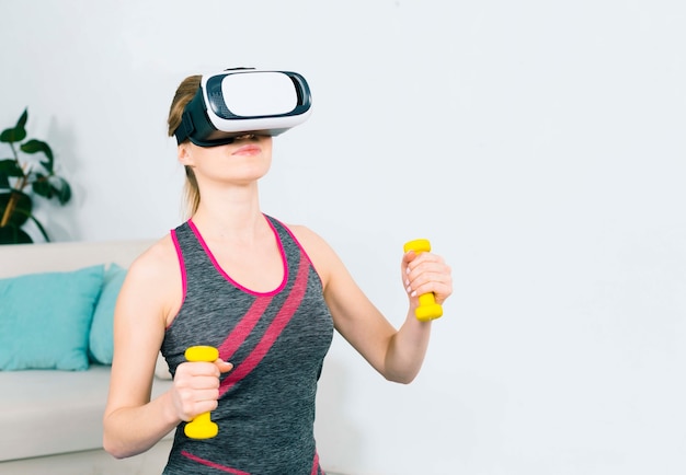 Primo piano della giovane donna che usando la cuffia avricolare di realtà virtuale che si esercita con i dumbbells gialli