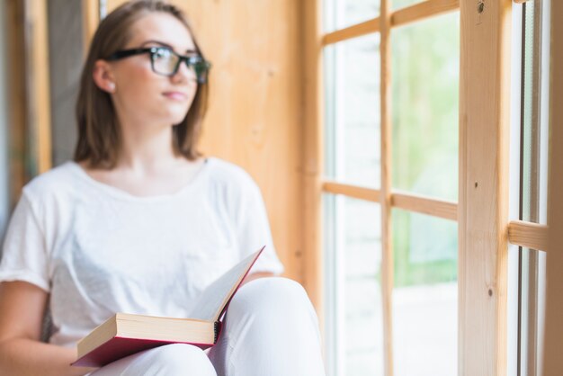 Primo piano della giovane donna con il libro che osserva fuori attraverso il vetro di finestra