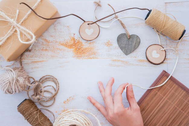 Primo piano della mano che fa la ghirlanda del cuore con la bobina e il contenitore di regalo avvolto sullo scrittorio bianco