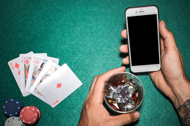 Primo piano della mano che tiene il cellulare e il bicchiere di whisky sul tavolo da poker