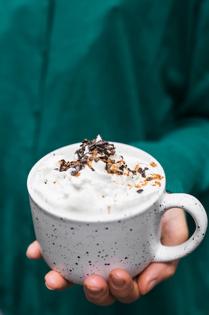 Primo piano della mano che tiene la tazza di cioccolata calda con panna montata e gocce di cioccolato